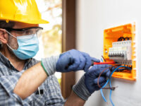 Electrician at work on an electrical panel protected by helmet, safety goggles and gloves; wear the surgical mask to prevent the spread of Coronavirus. Construction industry. Covid-19 Prevention.
